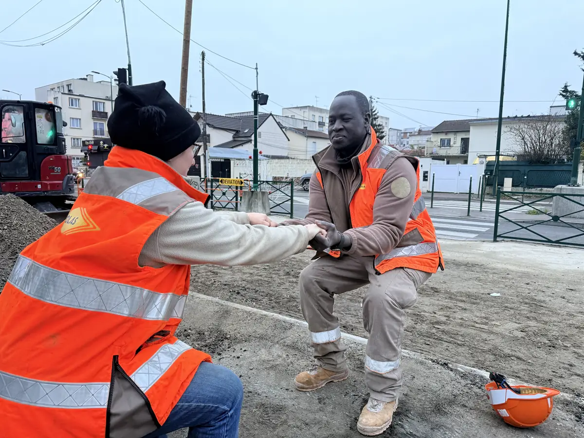 Un compagnon guide un collègue, genou au sol, par les mains, pendant un échauffement sur un chantier